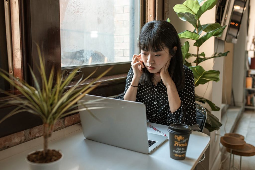 woman-in-black-and-white-long-sleeve-shirt-sitting-at-table-with-macbook-5915203 Asian woman focused on work in a modern office setting with laptop and coffee.