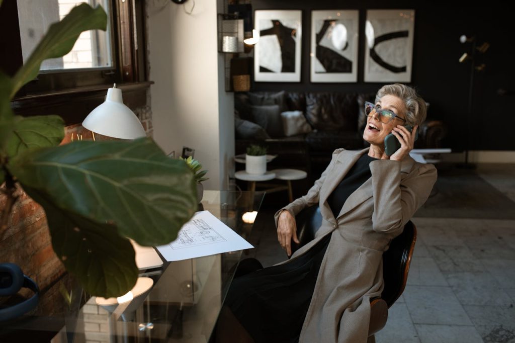 woman-in-brown-coat-holding-white-printer-paper-5921657 Senior woman in a modern office setting, smiling while on a phone call. Stylish and professional workspace.